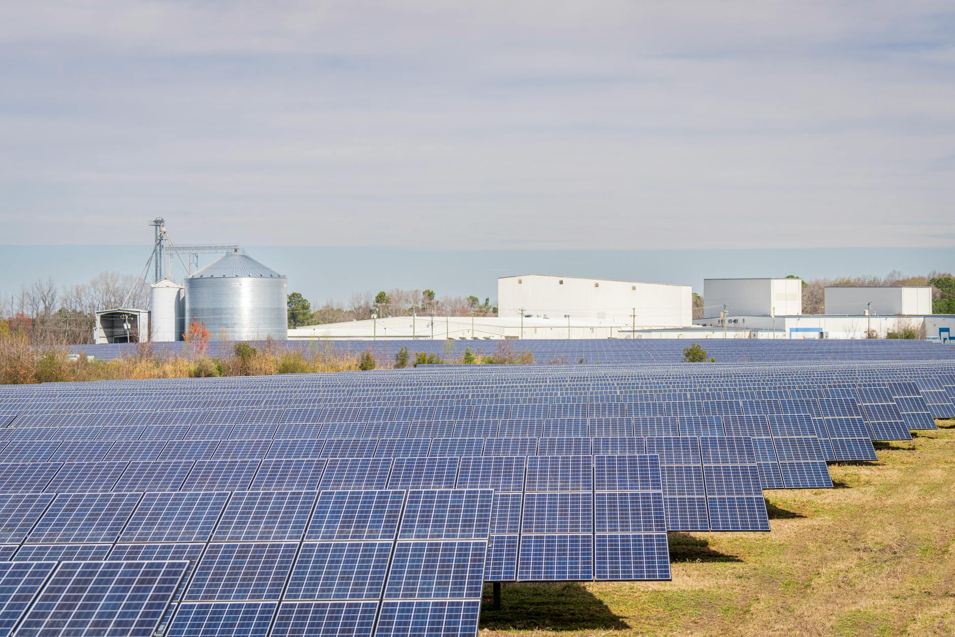 A large solar panel field with warehouses and silos in the background under a clear sky.