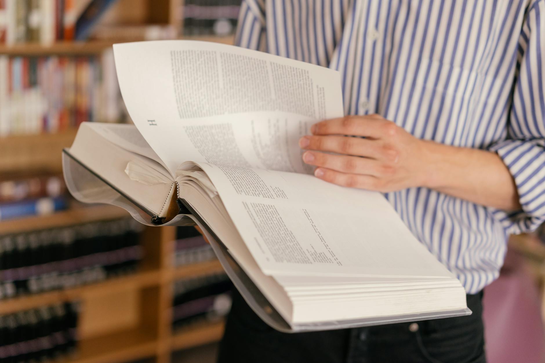 Close-up of a person reading a large open book in a library setting with bookshelves in the background.