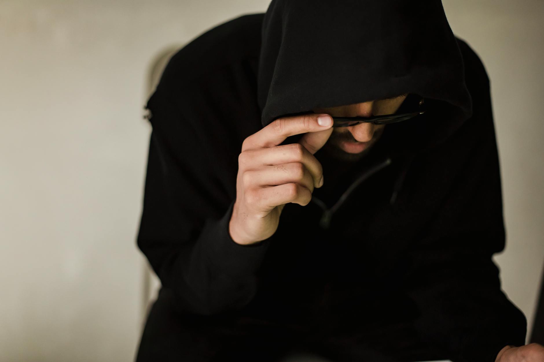 Crop focused male hacker covering head with hood while browsing laptop and lowering eyeglasses in surprise