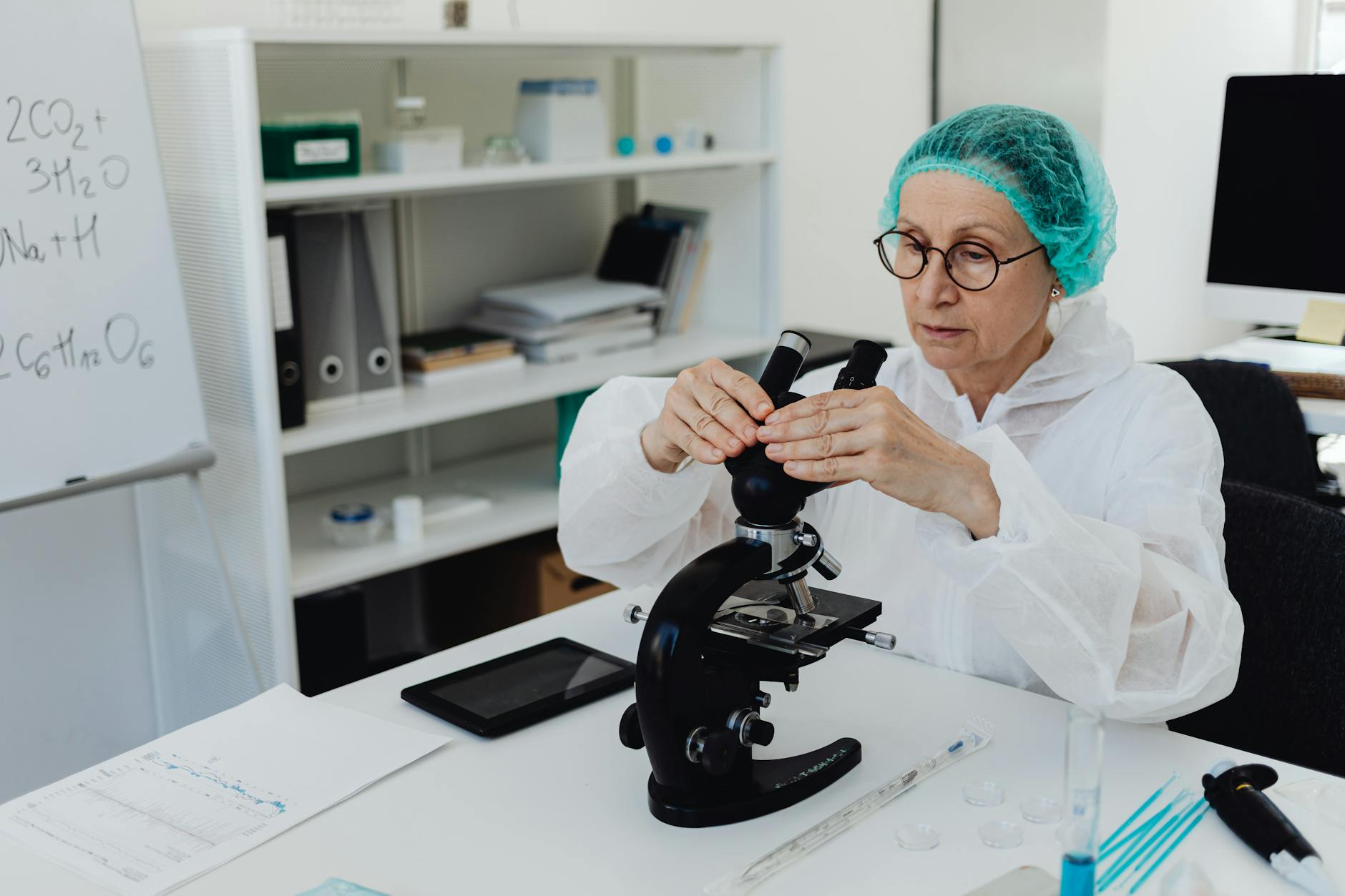 Elderly female scientist in PPE examining samples with a microscope in a modern lab.