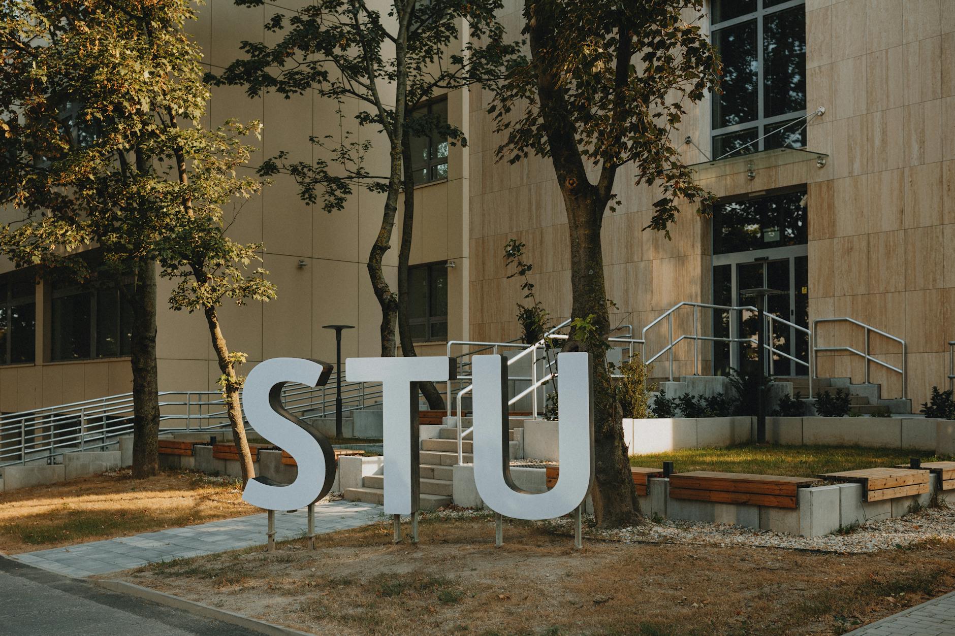 Modern building entrance with large STU letters surrounded by greenery and urban design.