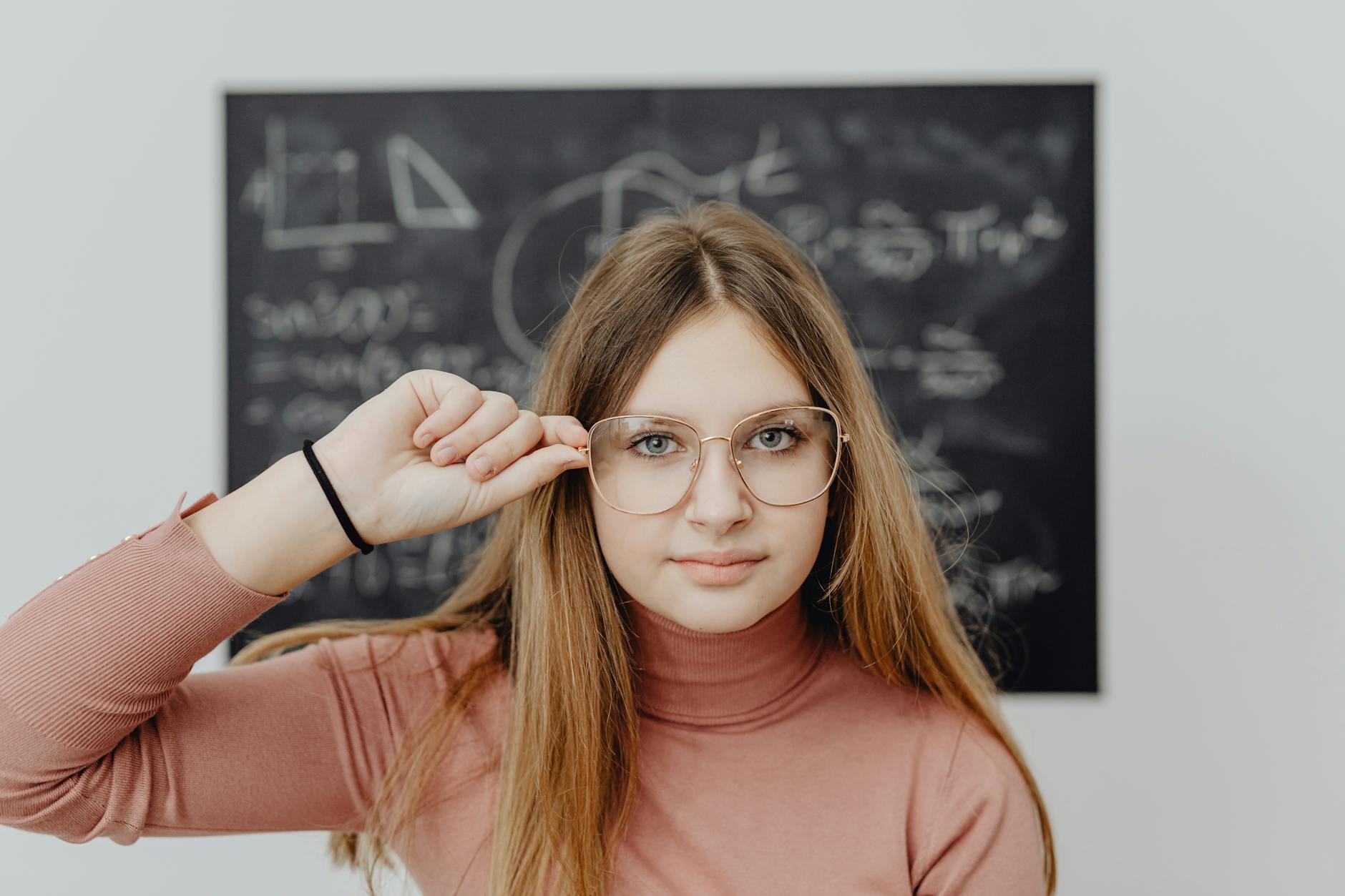 Portrait of a young woman wearing glasses in front of a chalkboard. Optimistic and thoughtful expression.