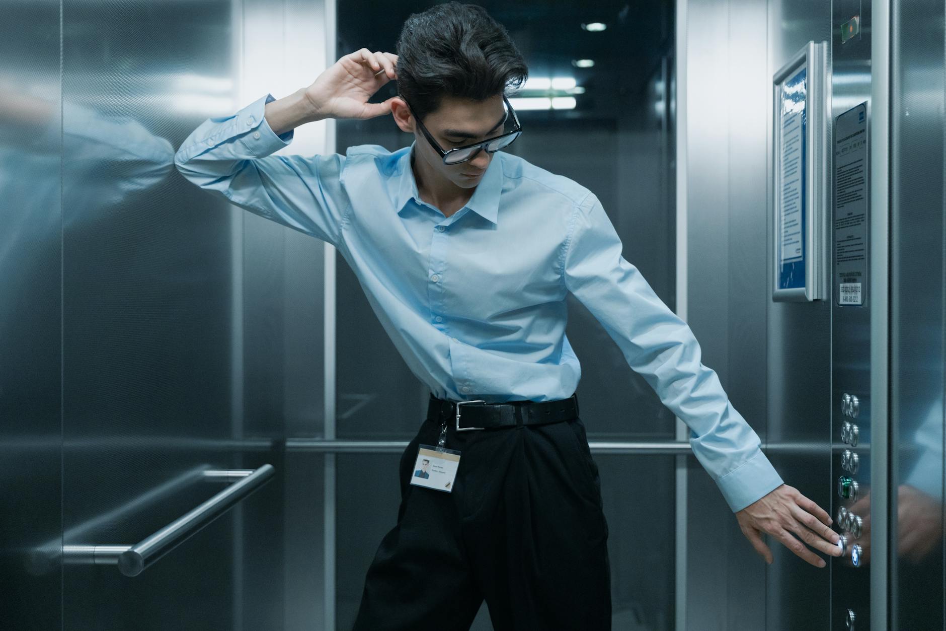 Stylish Asian man in office elevator adjusting his glasses, wearing professional attire.
