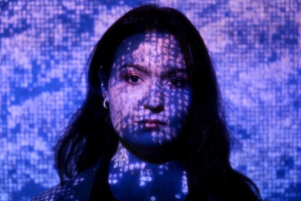 Close-up of a holographic human face fragmenting into digital pixels on a sleek transparent screen, ambient blue neon lighting, dark futuristic data center background, neural network visualization ove
