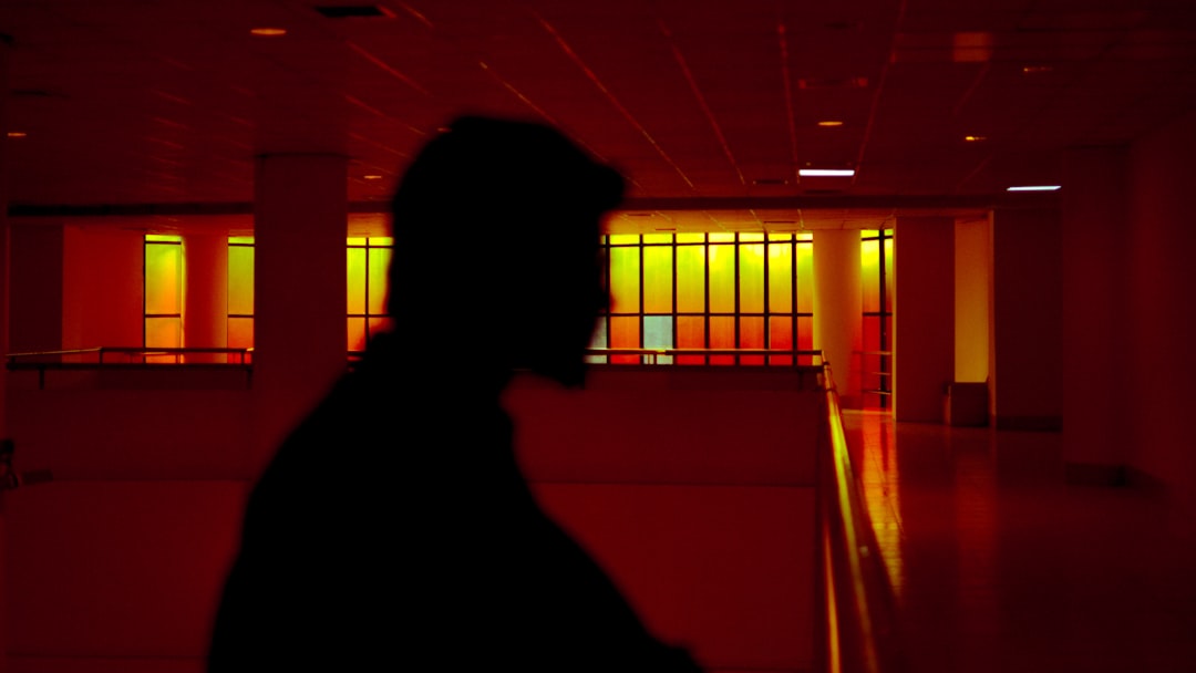Futuristic data center with ominous red ambient lighting, shadowed silhouette of engineer watching overwhelming server racks, neural network visualization glowing on holographic display, crisis atmosp