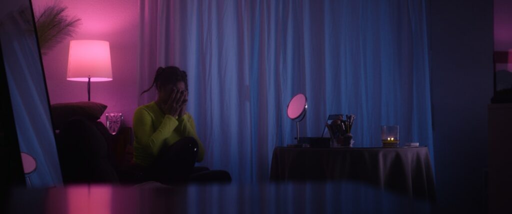 Young person sitting alone in dimly lit modern apartment, glowing smartphone illuminating their face, holographic AI interface floating nearby, cool blue ambient lighting, futuristic loneliness concep