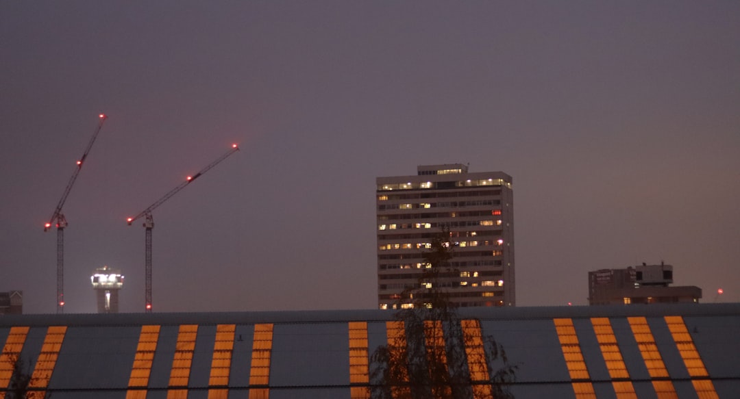 Aerial view of massive industrial data center complex at dusk, construction cranes beside operational buildings, contrast between glossy marketing billboard and gritty reality, dramatic sky, professio