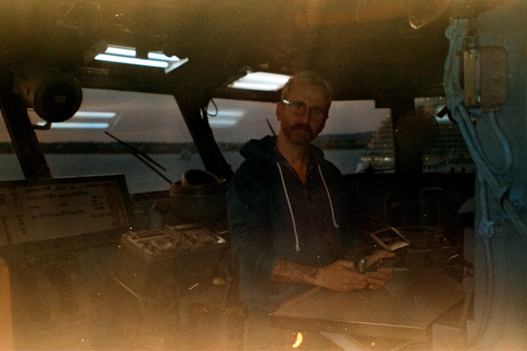 Overwhelmed technician at outdated maritime radar console with red warning lights, cluttered screens showing false alerts, dark control room, chaotic atmosphere, failure of legacy systems