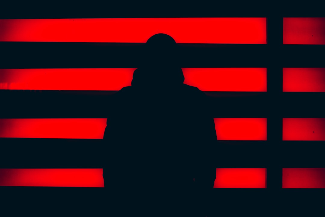 Dark moody AI ethics concept with shadowed human figure facing glowing red neural network, cybersecurity dashboard reflections, dramatic contrast lighting, abstract warning data patterns, professional