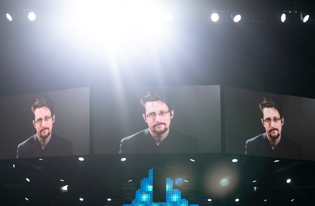 Dynamic wide-angle of speaker on stage with massive LED screen showing data analytics, dramatic spotlighting, engaged audience silhouettes, corporate tech event, purple and cyan ambient glow