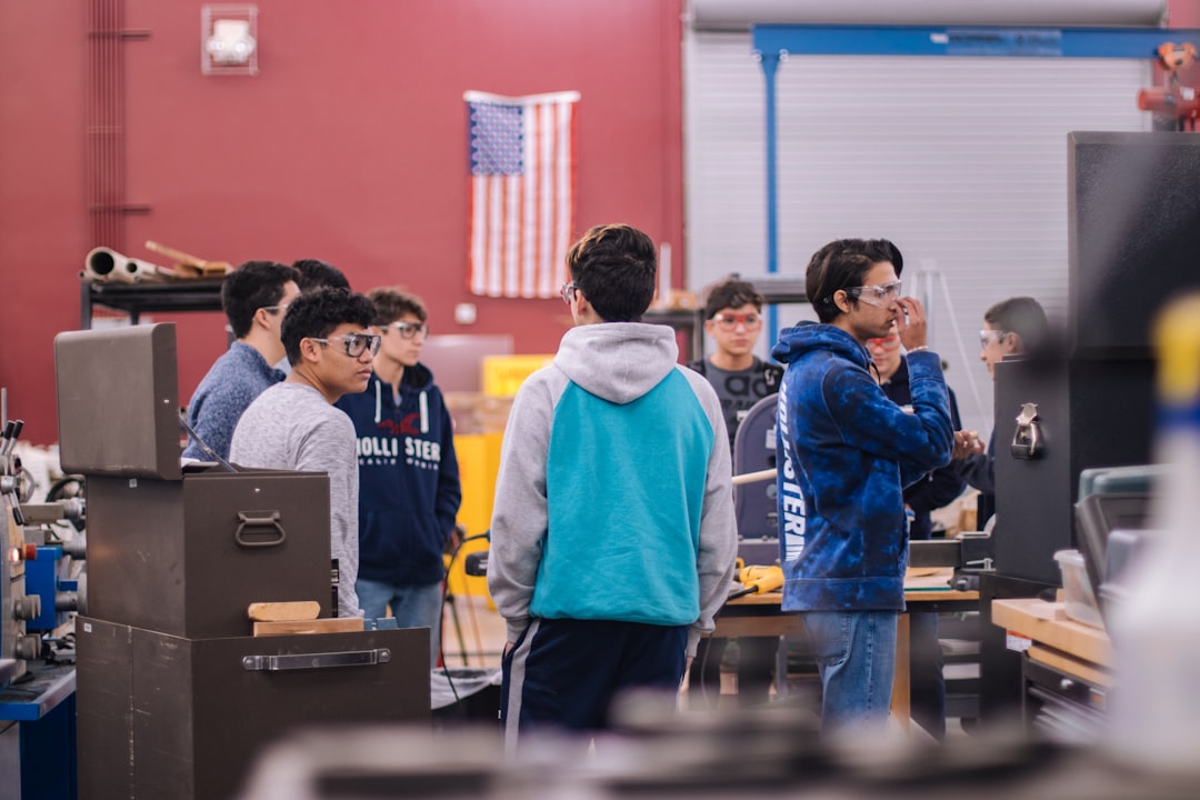 Diverse students and young workers facing away from abandoned classroom toward automated robotics assembly line through glass wall with dramatic overhead lighting
