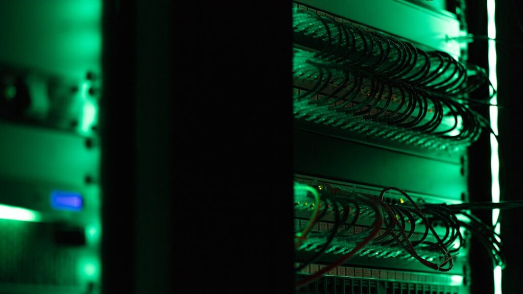 Futuristic server room with flickering red warning lights, exhausted technician in hard hat examining overheating server racks, dark moody ambient lighting, collapsing infrastructure, professional tec