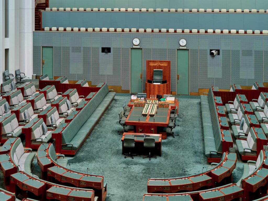 Futuristic legislative chamber with holographic AI regulation displays, ambient blue lighting, suited professionals reviewing digital policy documents, sleek modern government setting, neural network