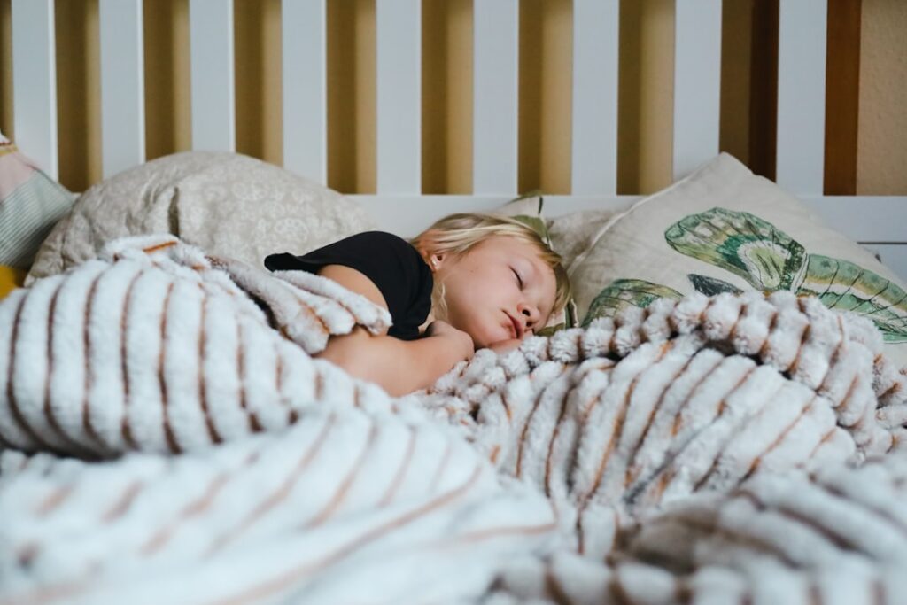 Young child sleeping peacefully in futuristic bedroom with soft blue ambient lighting, holographic AI assistant hovering nearby, sleek modern design, neural network patterns projected on walls