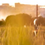 Tropical farmer examining drought-stressed crops under harsh sun, split composition with holographic weather data overlay, warm amber and deep green tones, cinematic documentary style