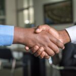 Two business professionals shaking hands in a futuristic data center with holographic displays showing neural networks, ambient blue lighting, sleek server racks in background, cinematic tech aestheti