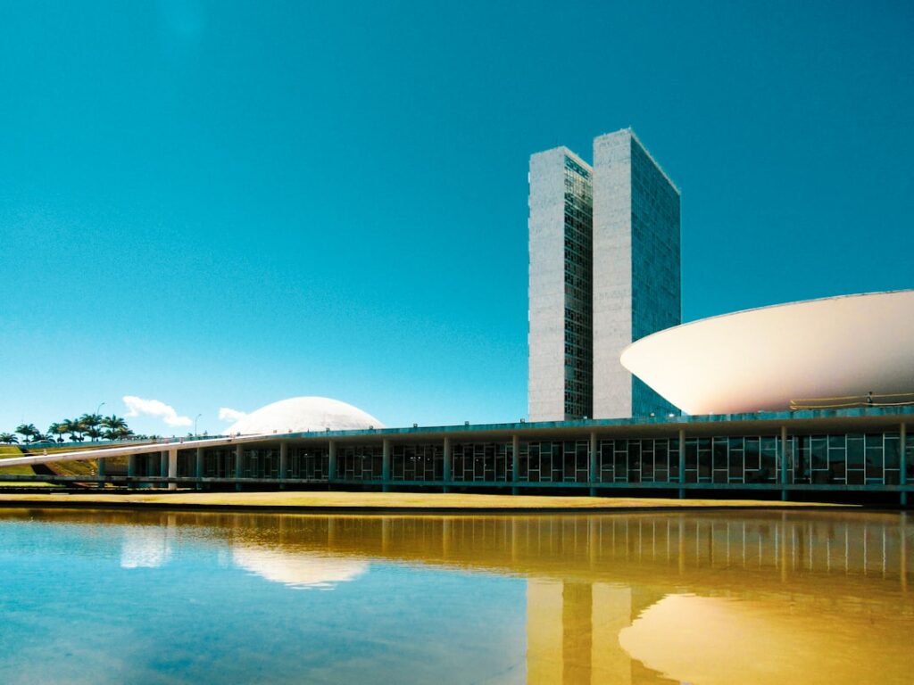 Futuristic Brazilian courthouse exterior at golden hour, holographic data streams overlaying classical columns, silhouetted professionals ascending steps, sleek ambient lighting, teal and amber tones,