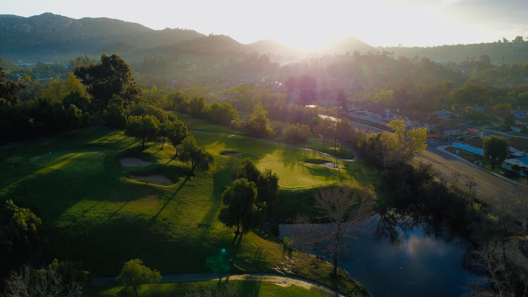 Aerial view of lush golf course at golden hour with holographic data overlays, financial charts floating in sky, professional investor silhouette observing, sleek futuristic interface elements, ambien