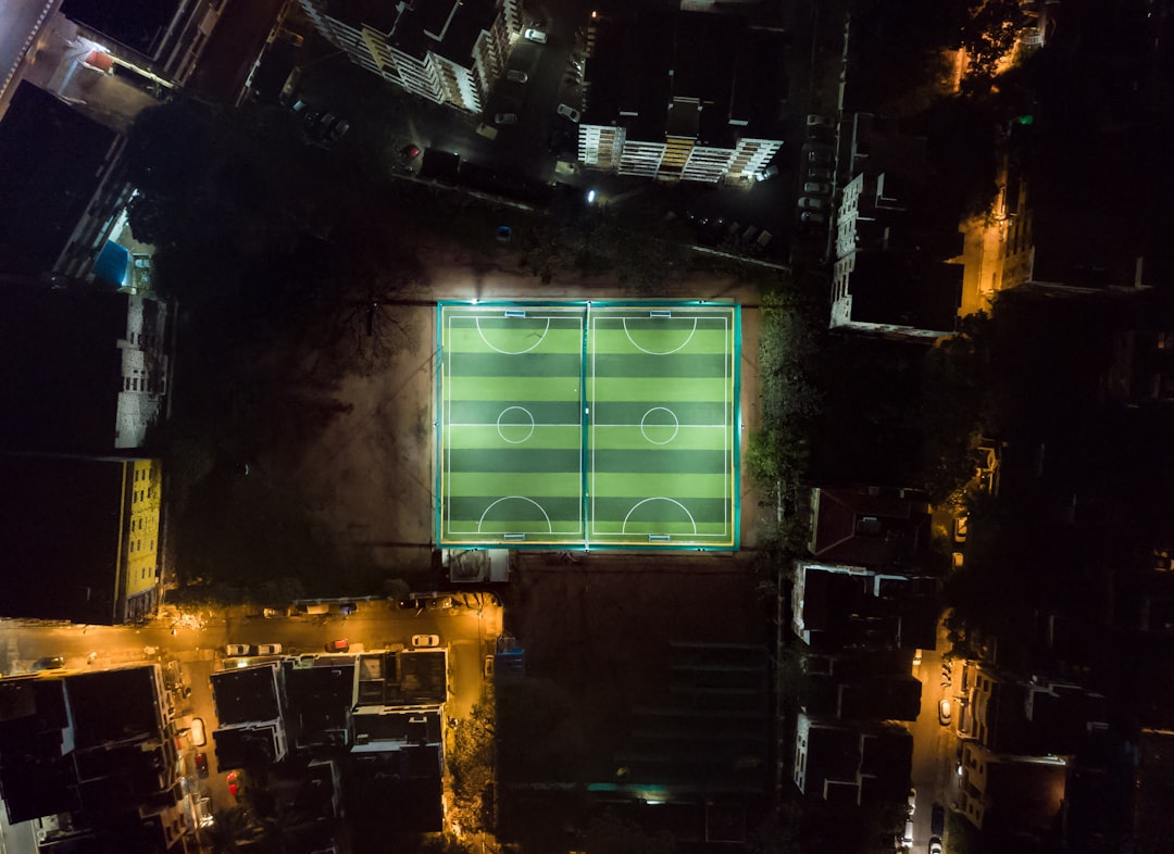 Aerial view of a sleek modern soccer stadium at dusk with holographic tactical overlays and neural network visualization floating above the pitch, professional sports technology concept, ambient blue