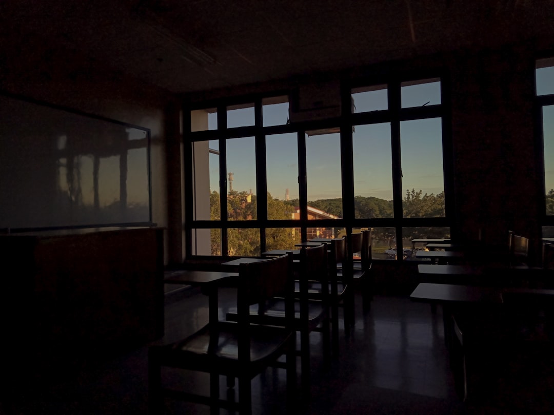 Empty classroom with abandoned coding stations, holographic training modules glitching, neural network visualization fading, cool desaturated tones, broken pipeline metaphor