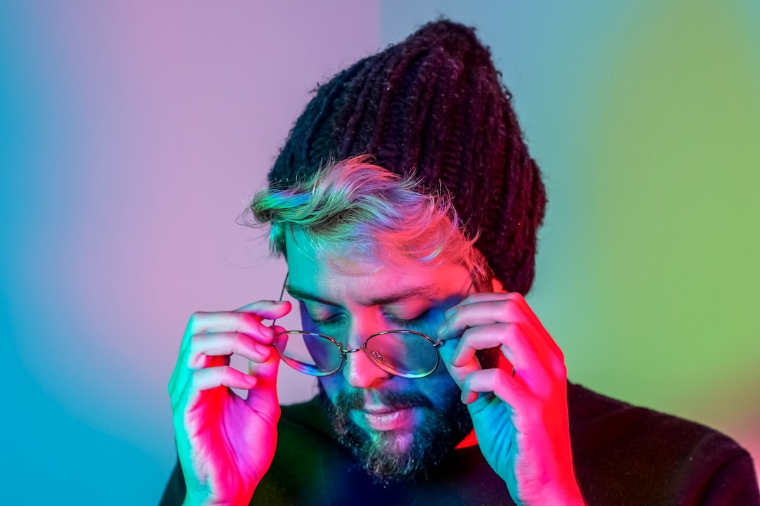 Exhausted young developer removing glasses while surrounded by multiple glowing screens showing neural network anxiety patterns in moody neon-lit workspace