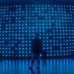 Futuristic human silhouette pausing before a massive holographic globe display, cool blue ambient lighting, sleek glass architecture, contemplative mood, neural network patterns floating in background