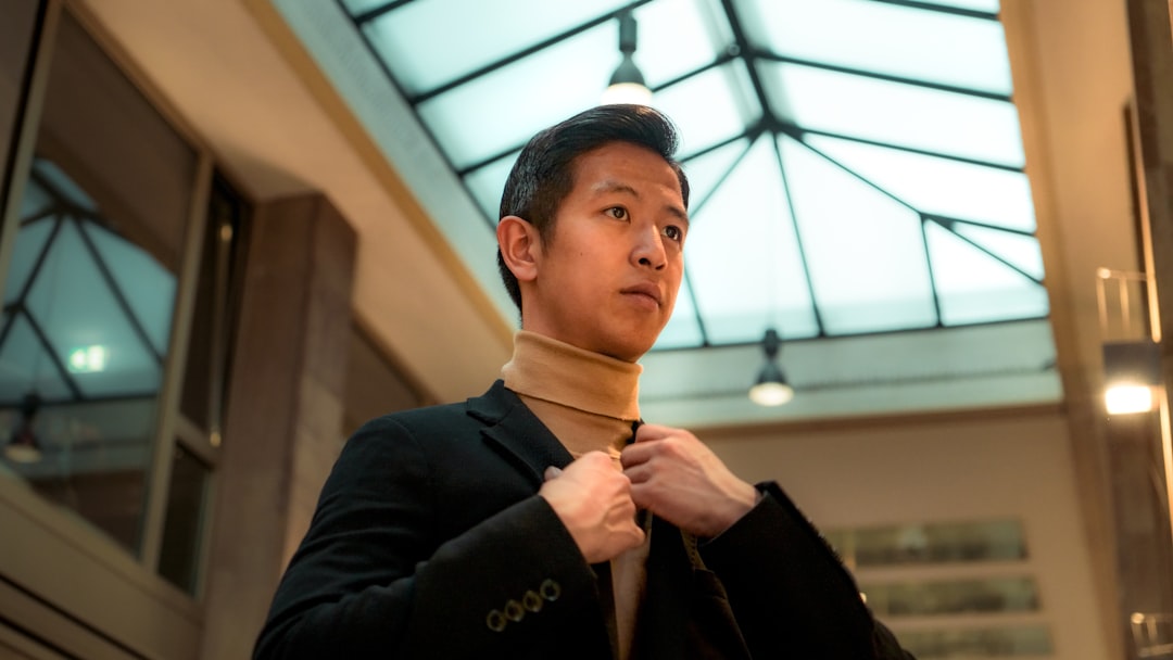 Young Asian tech CEO in sleek black turtleneck, dramatic side lighting, standing before floor-to-ceiling glass windows overlooking futuristic city skyline, holographic data projections floating around